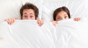 Funny couple in bed looking and peeking over sheets surprised. Young interracial couple, Asian woman, Caucasian man.